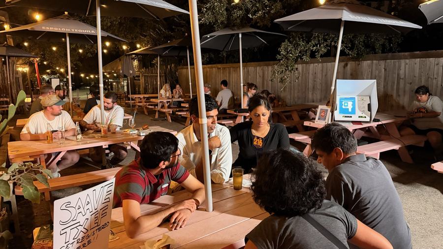 Neighbors at picnic tables on a bar patio during a voter registration trivia night, with a demo voting machine set up nearby